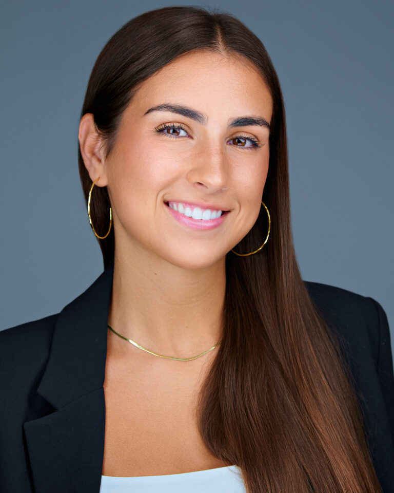Professional headshot of Cassie Arietta, a young woman with long brown hair, wearing gold hoop earrings, a gold necklace, and a black blazer over a light top, smiling against a soft blue-gray background.