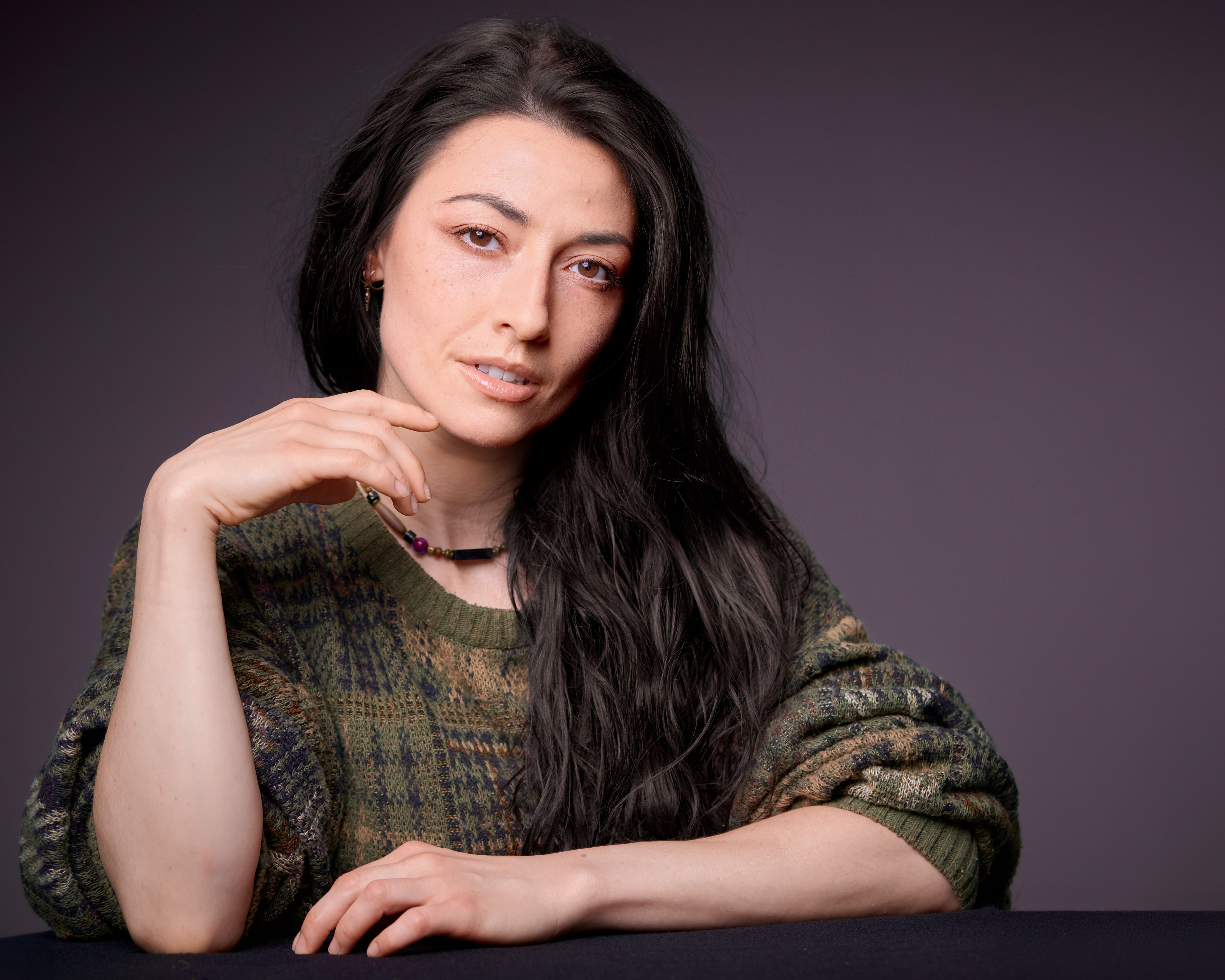 Professional woman with long dark hair resting hand near chin, wearing a green patterned sweater, posed against a dark background.