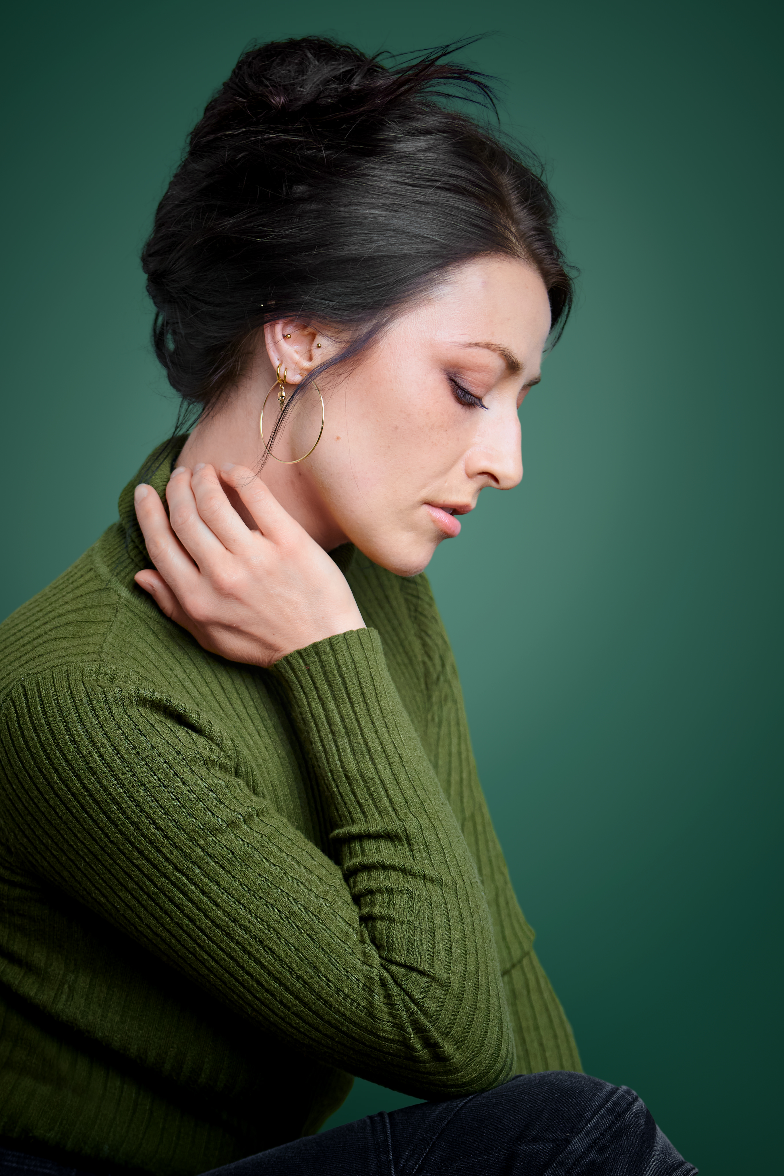 Professional woman in profile wearing hoop earrings and green ribbed sweater, looking downward in a serene pose.