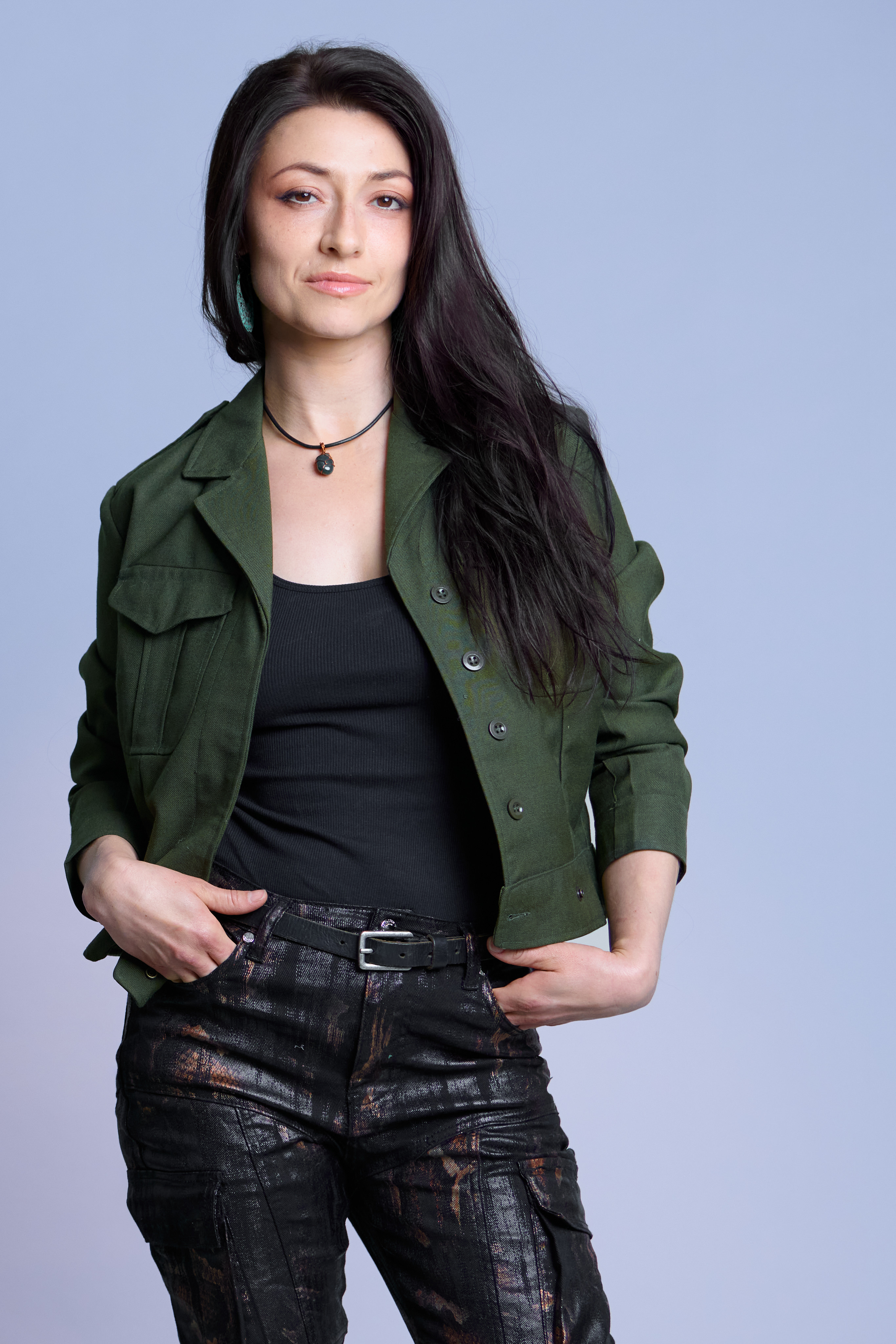 Professional woman standing with confident posture, wearing green jacket and black top, against light blue background.
