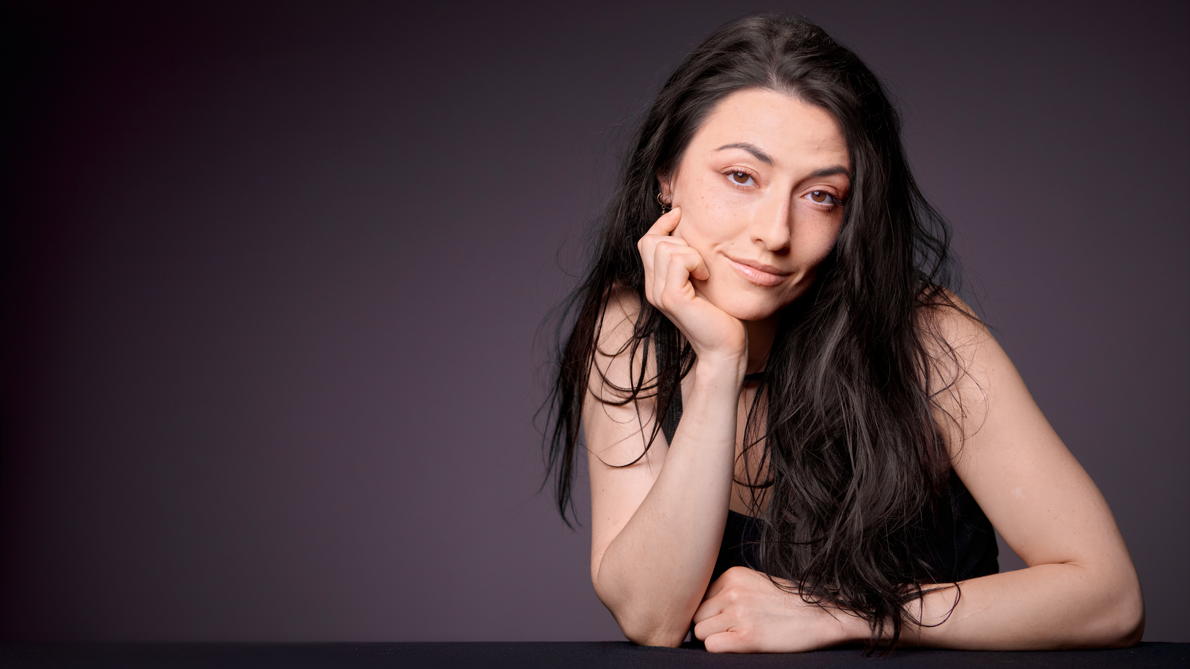 Professional headshot of a woman with long dark hair, chin resting on hand, seated at a table, right of frame on a dark plum background.