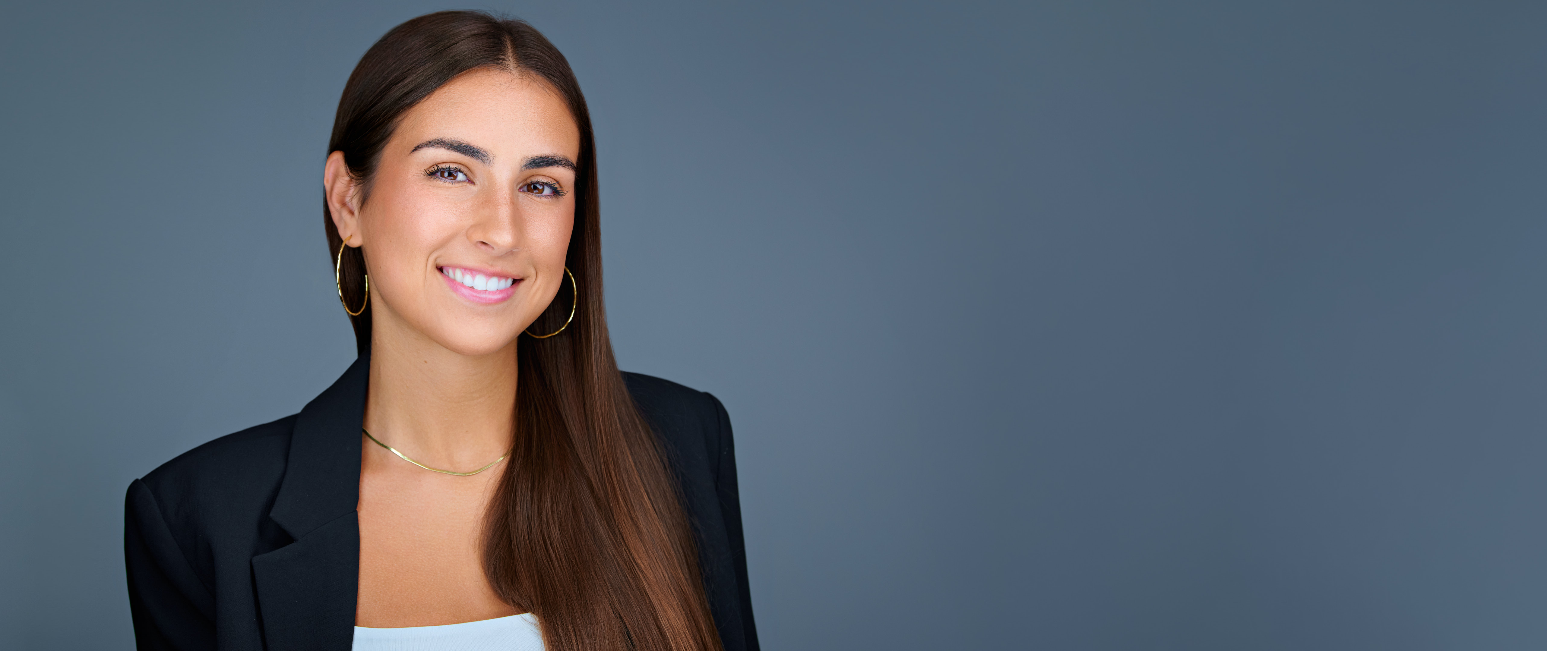 A polished, professional headshot of Cassie Arietta, captured with a clean blue-gray backdrop. She is wearing gold hoop earrings, a delicate gold necklace, and a black blazer layered over a light top. Her straight, long brown hair falls over her shoulder, and she smiles warmly at the camera, conveying approachability and confidence.