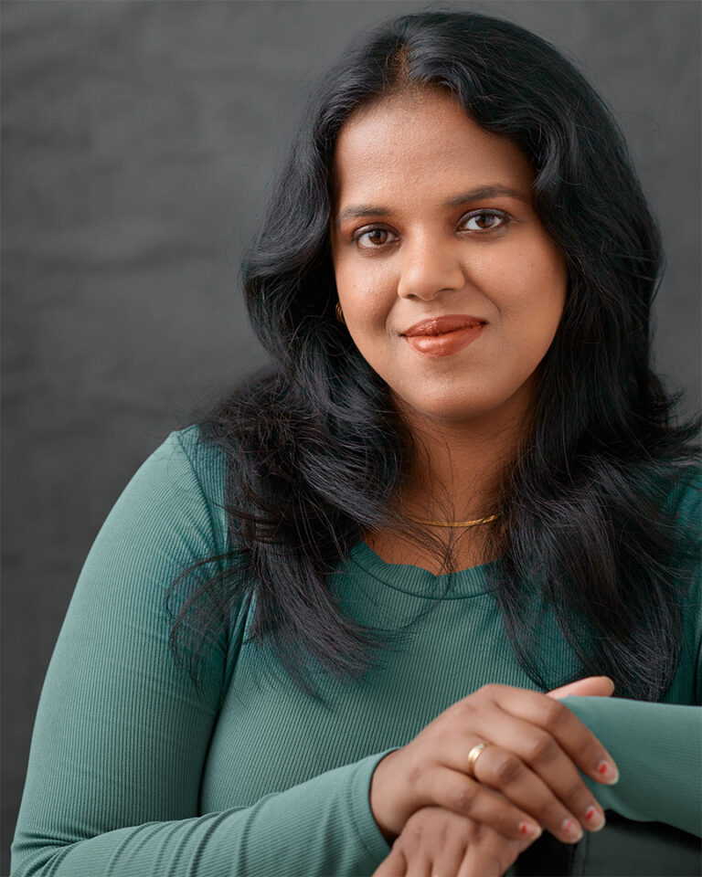 Woman with long dark hair in a soft green top, gently smiling with arms crossed, photographed against a neutral gray backdrop with soft, even light.
