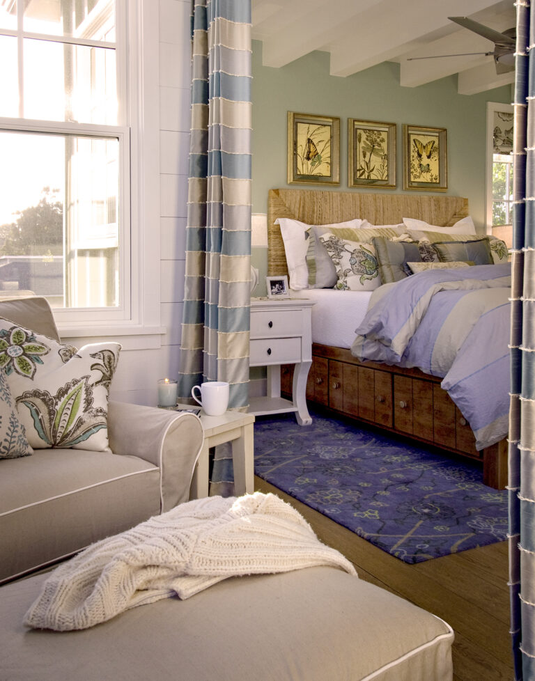 Sunlit bedroom with a blue‑and‑sage palette—striped curtains, rustic wood headboard beneath three botanical prints, blue rug, and a beige armchair with floral pillow and knitted throw.