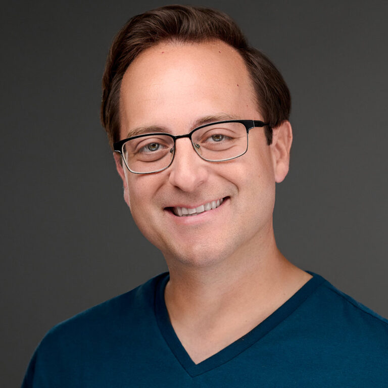 Professional portrait of a man in blue V-neck shirt, neutral backdrop.