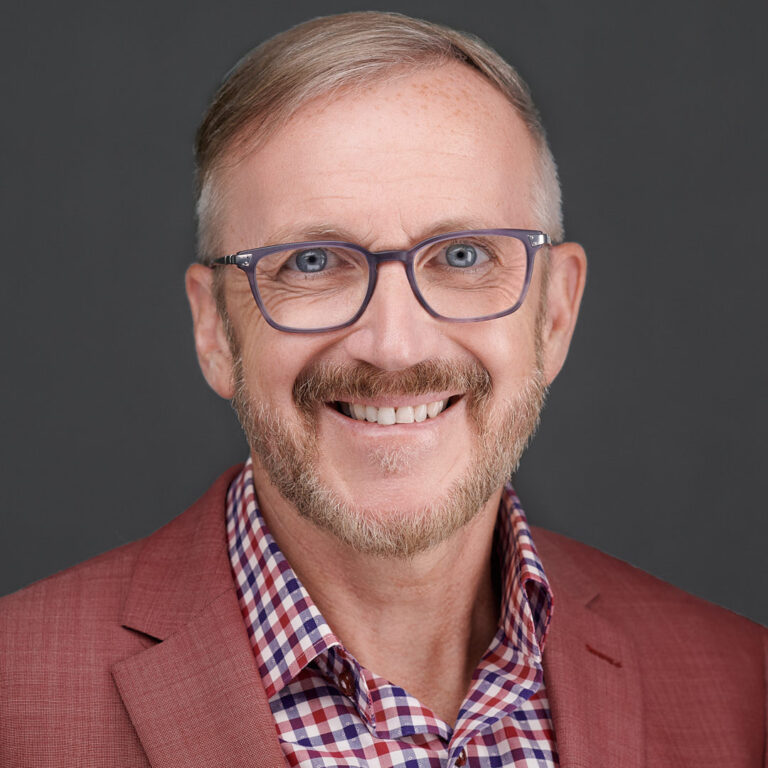 Corporate portrait of a man in red blazer and checked shirt, dark background.