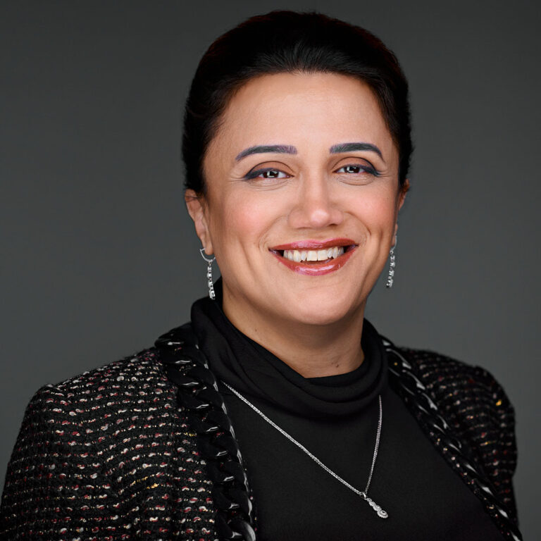 Corporate headshot of a woman in black jacket with subtle jewelry, gray background.