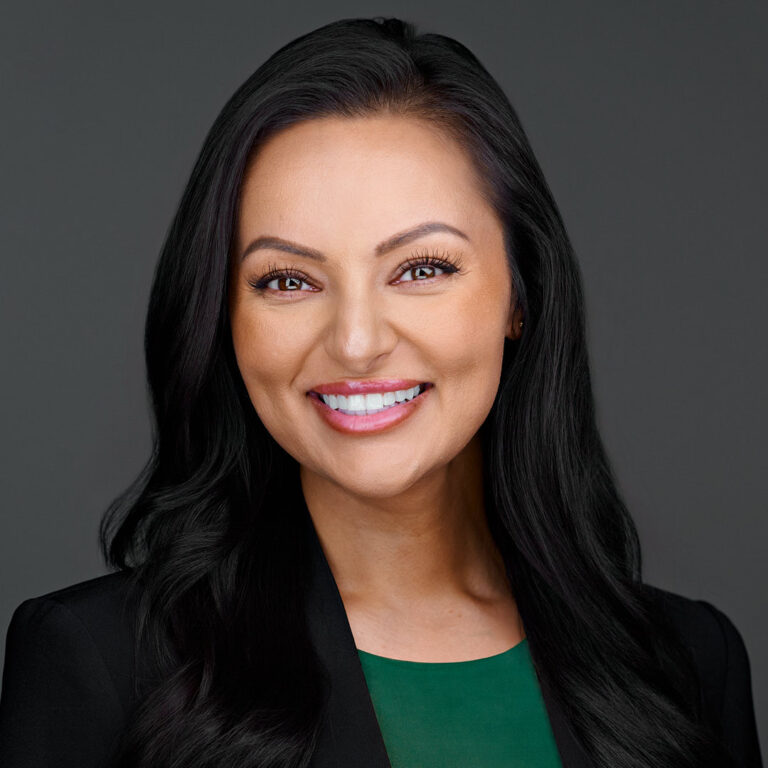 Professional headshot of a woman in black blazer with green top, dark background.