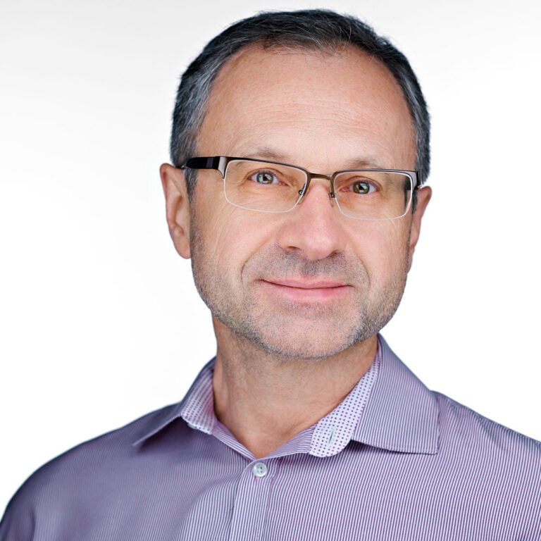Corporate headshot of a man in light purple shirt and glasses, bright background.