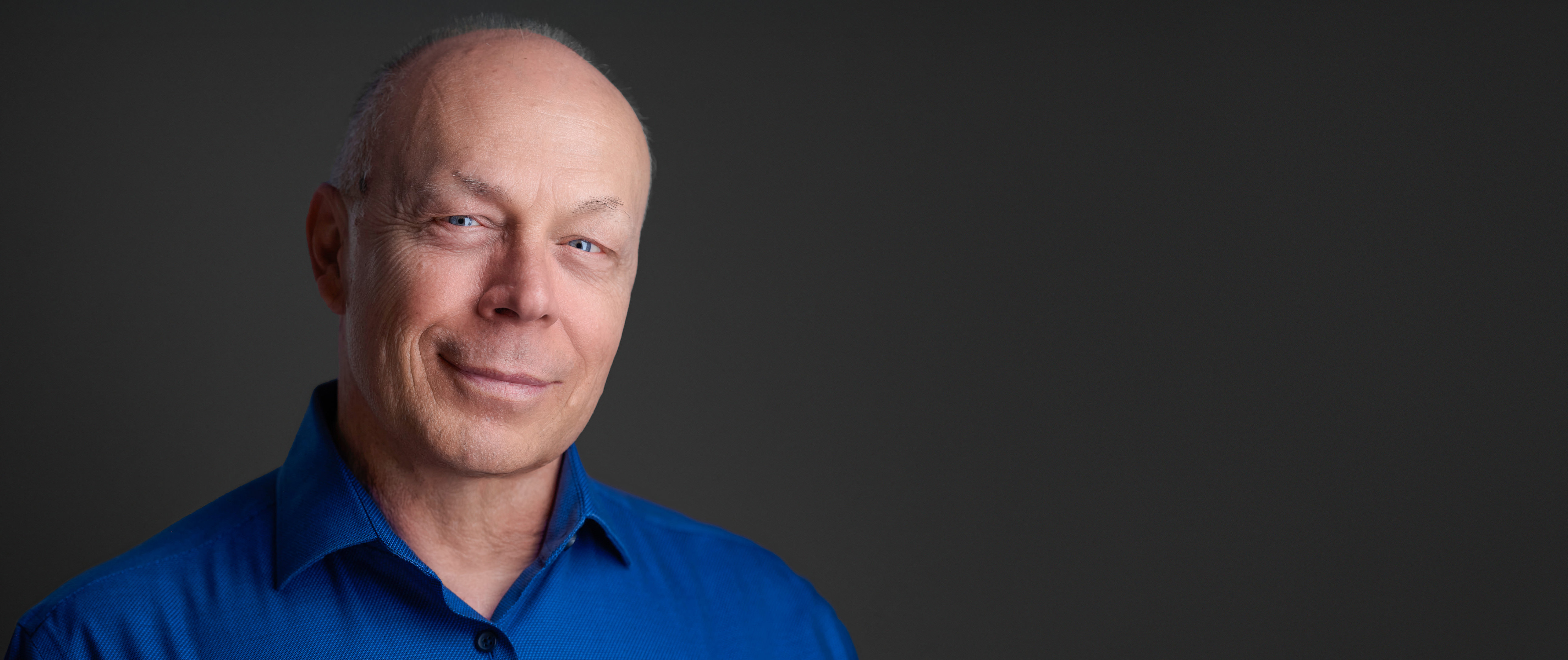 Professional portrait of photographer Dean Birinyi, smiling confidently in a blue shirt against a dark gray background.