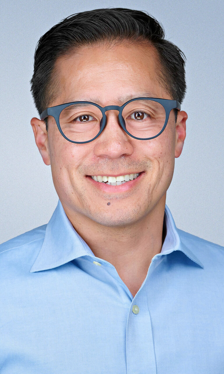 Professional corporate headshot of a smiling male executive wearing blue glasses and a light blue dress shirt, photographed by Dean Birinyi Photography in Palo Alto, California. Image used on the company headshot system page to represent polished, brand-aligned portraits for Bay Area teams.