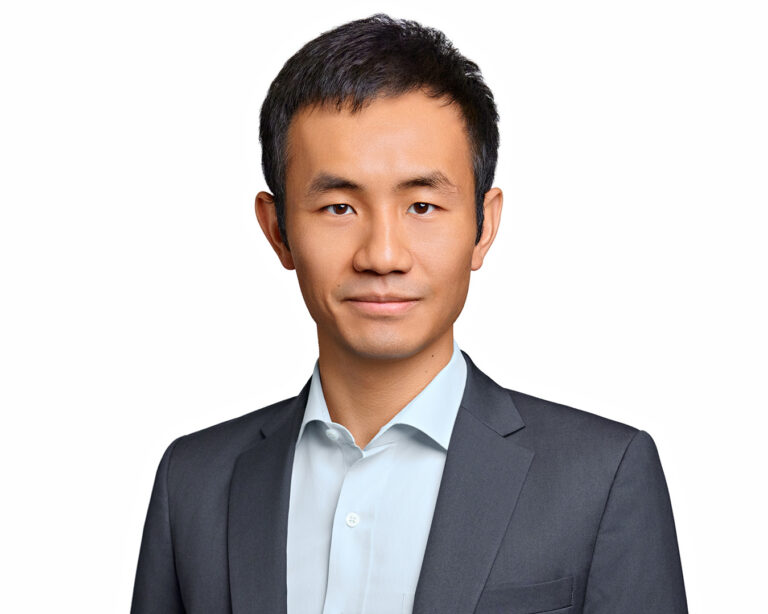 Professional headshot of a man in a gray blazer and blue shirt, photographed with clean studio lighting on a white background.