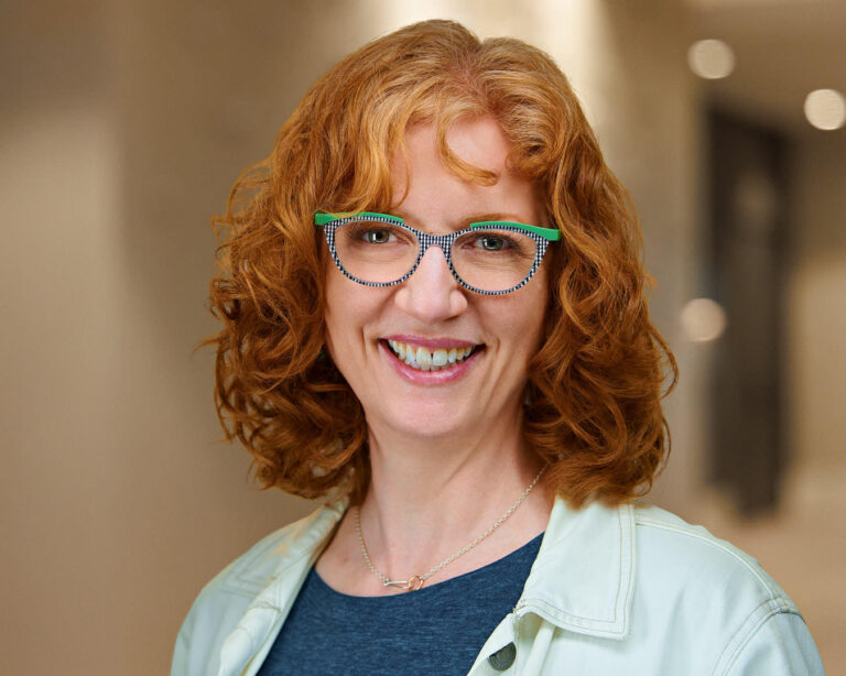 On-location corporate headshot of a professional woman with red curly hair and glasses, photographed in San Mateo, CA office environment.