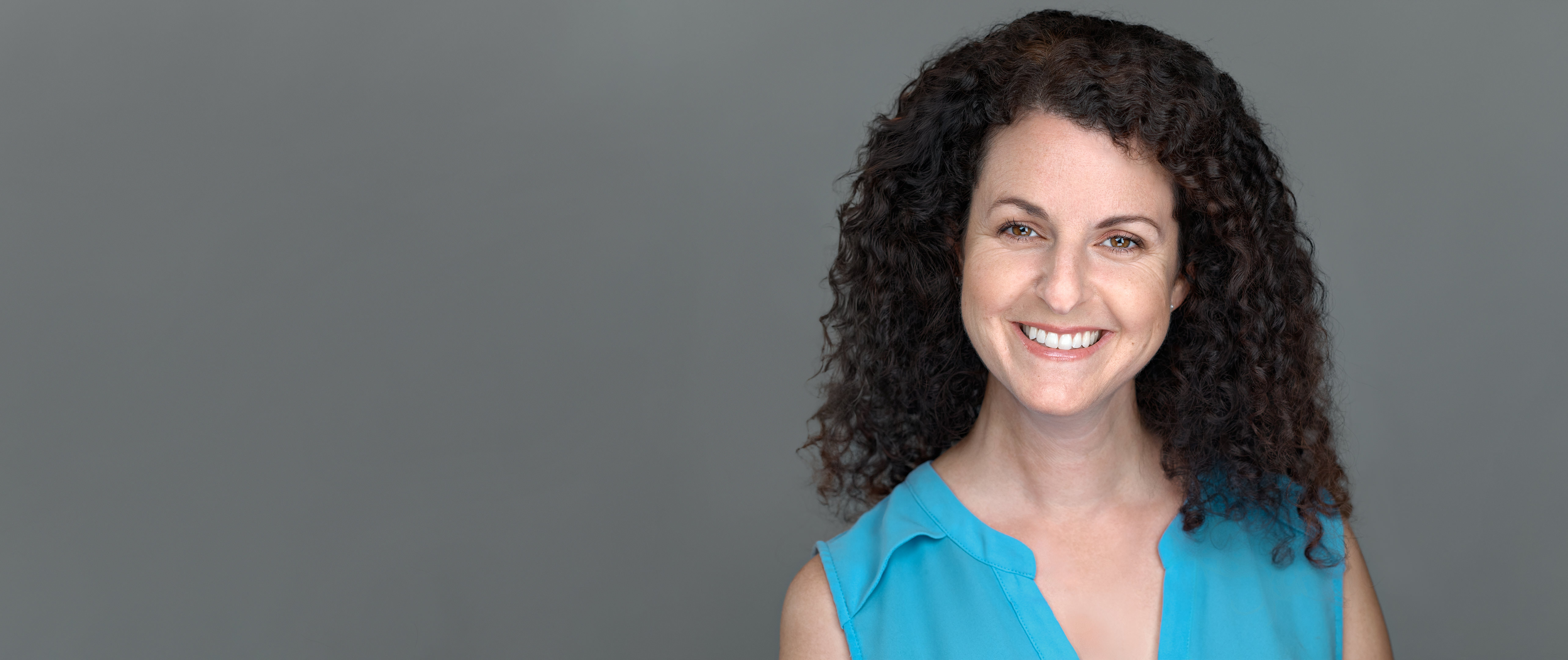 On-location corporate headshot of a professional woman with curly dark hair wearing a teal blouse, photographed in San Mateo, CA.