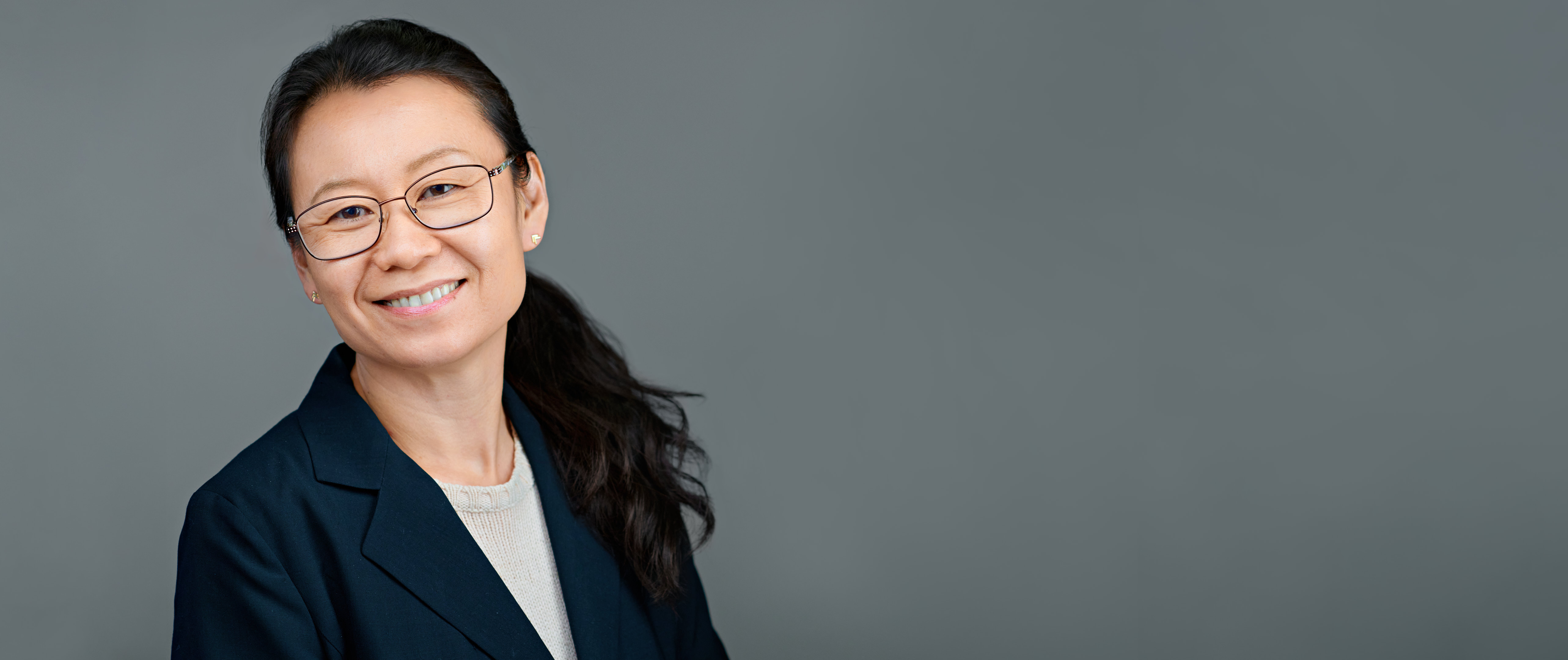 Professional company headshot of a smiling woman in glasses and a navy blazer against a gray background.