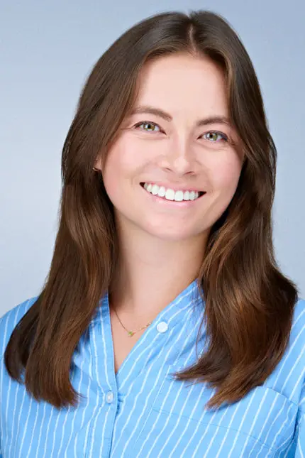 Professional corporate headshot of a smiling woman with long brown hair wearing a light blue striped shirt against a light gray background.