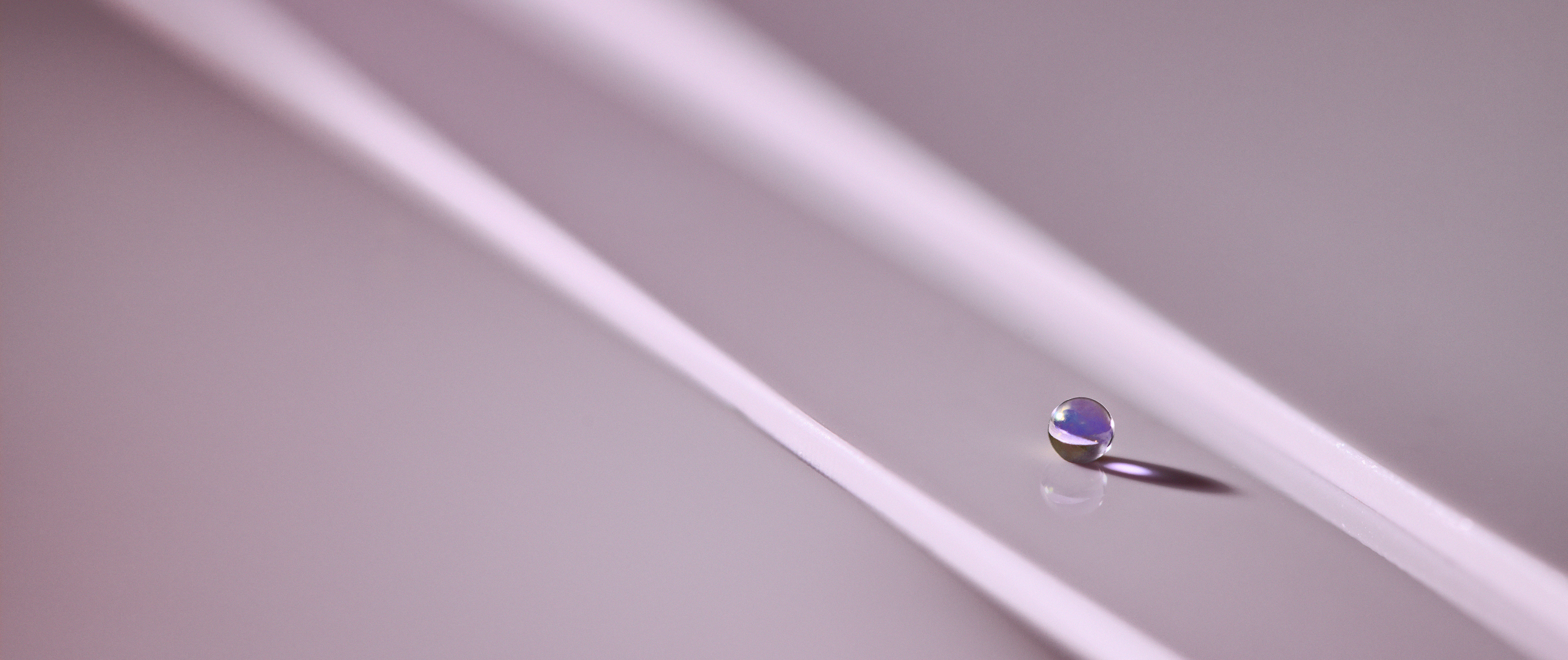 Abstract macro photograph of a translucent bead resting between two pale diagonal lines on a violet surface, casting a reflection and elongated shadow.