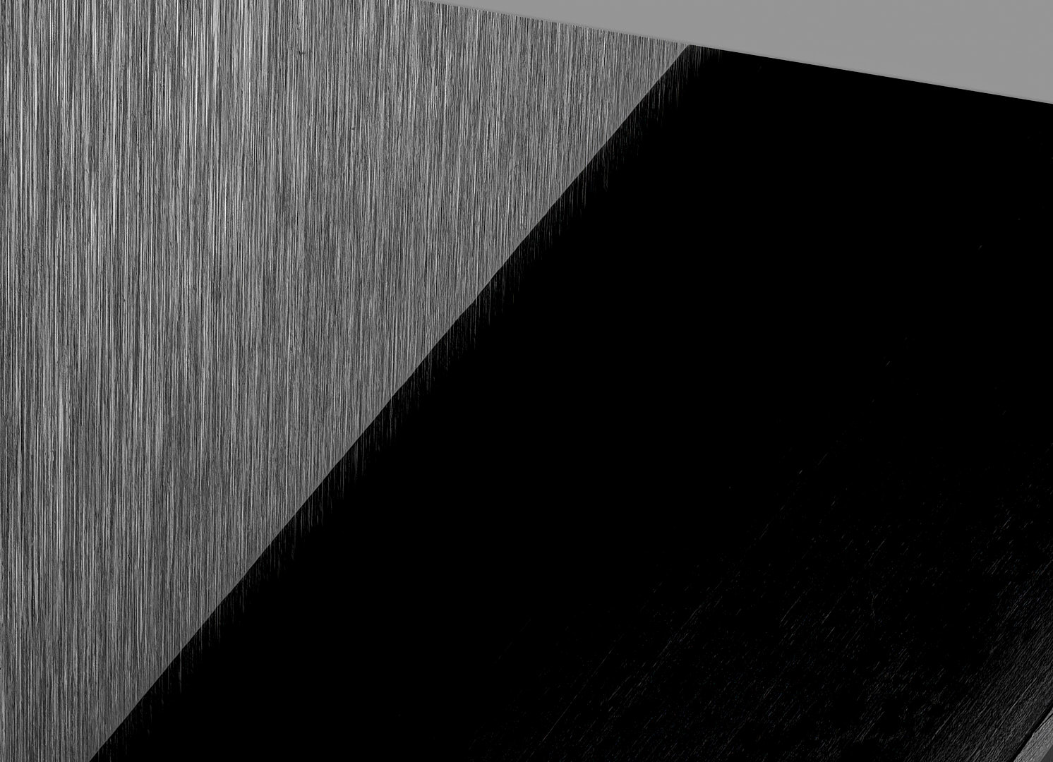 Abstract monochrome photograph divided diagonally between a finely textured light surface and a large featureless black area, with a narrow dark edge and faint material detail visible along the lower border.
