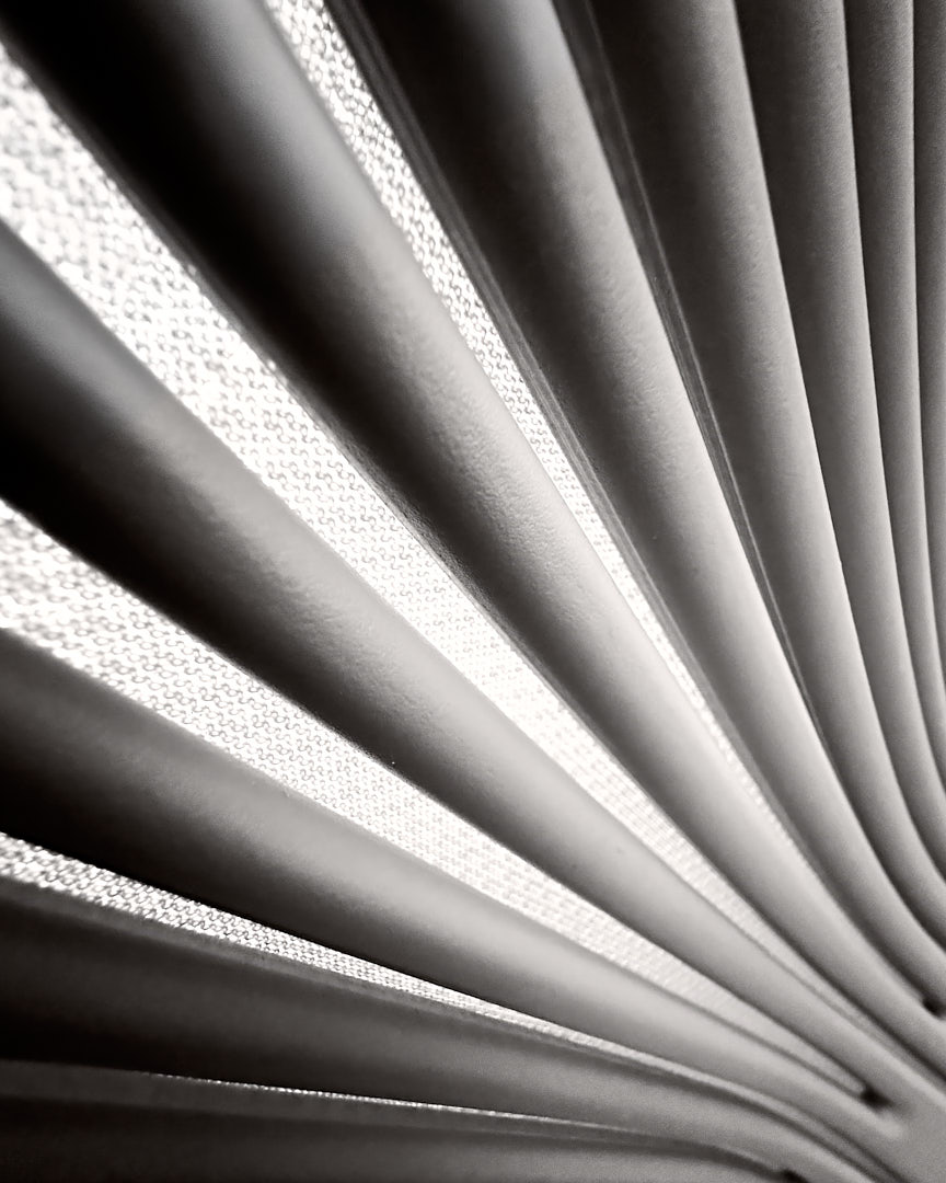 Black-and-white abstract photograph of the back of a conference-room chair, with repeated curved bands and bright textured openings converging toward the lower right.