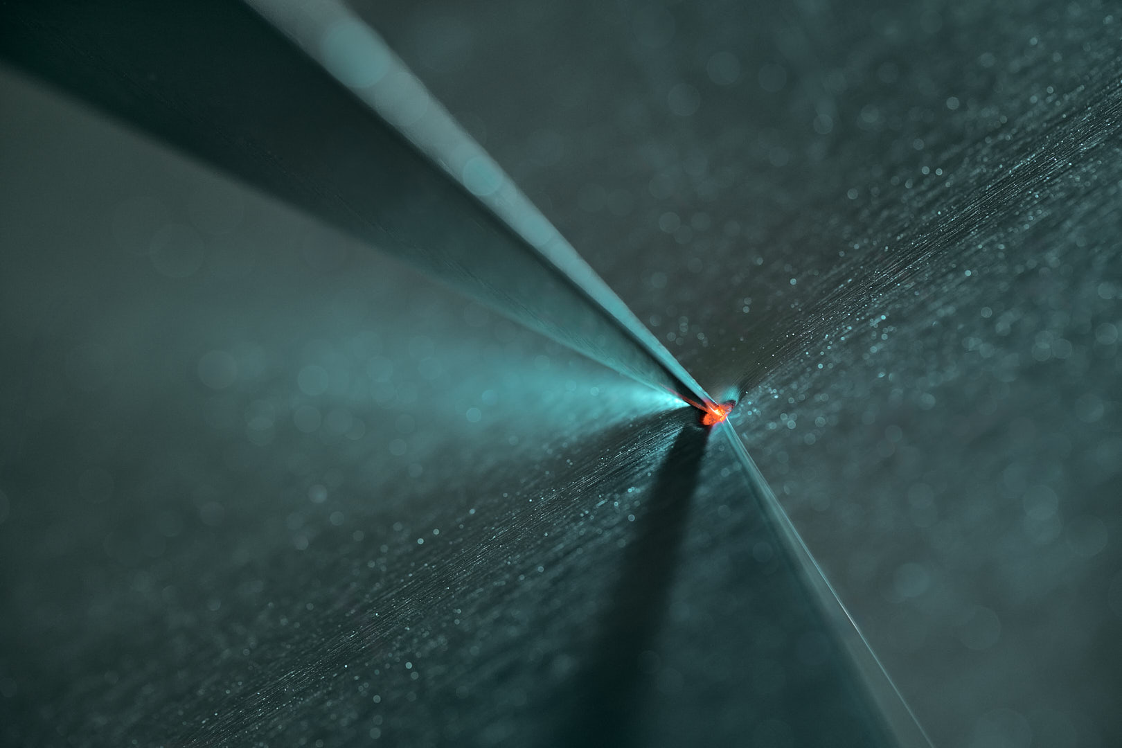 Abstract horizontal photograph of dark blue and teal angular planes converging on a small bright white point near center-right, surrounded by red-orange color and thin cyan highlights at a sharp break in the surface.