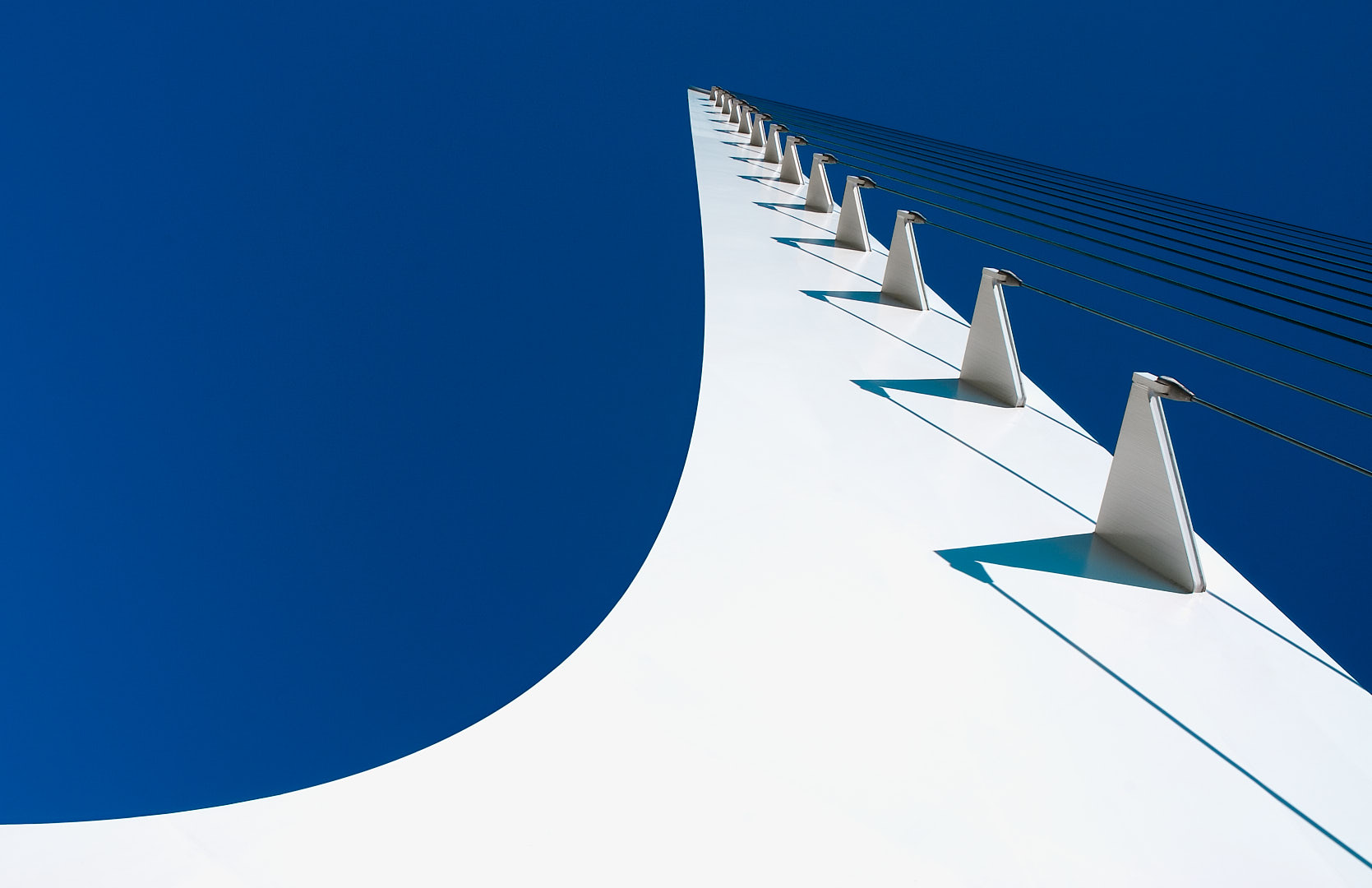Upward view of the Sundial Bridge showing a sweeping white tower, repeating cable mounts and shadows, and taut cables against a deep blue sky.