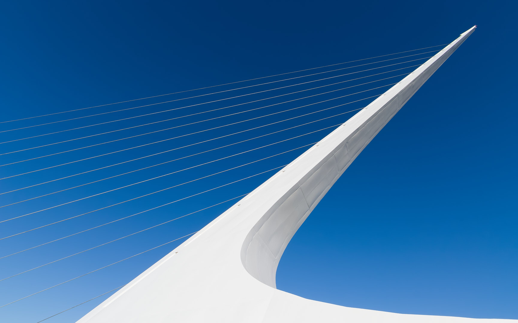 Upward view of the Sundial Bridge showing a sweeping white tower and radiating suspension cables against a deep blue sky.