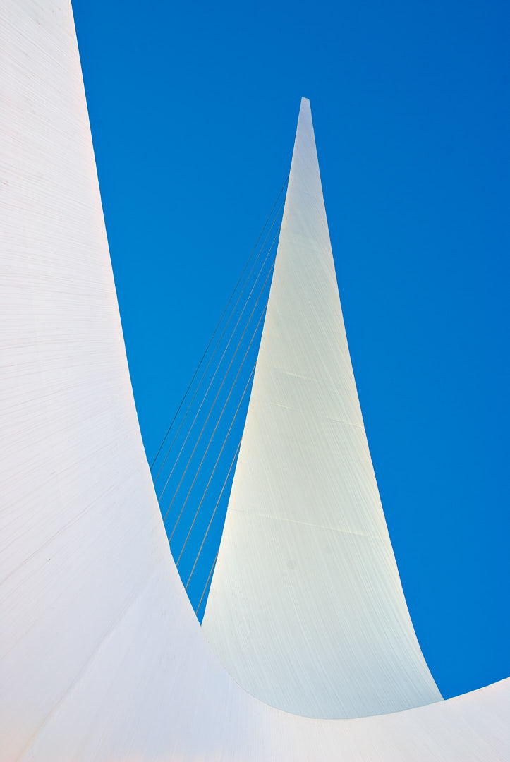 Low-angle view beneath the Sundial Bridge showing two sweeping white structural forms, a narrow central tower, and suspension cables against a vivid blue sky.