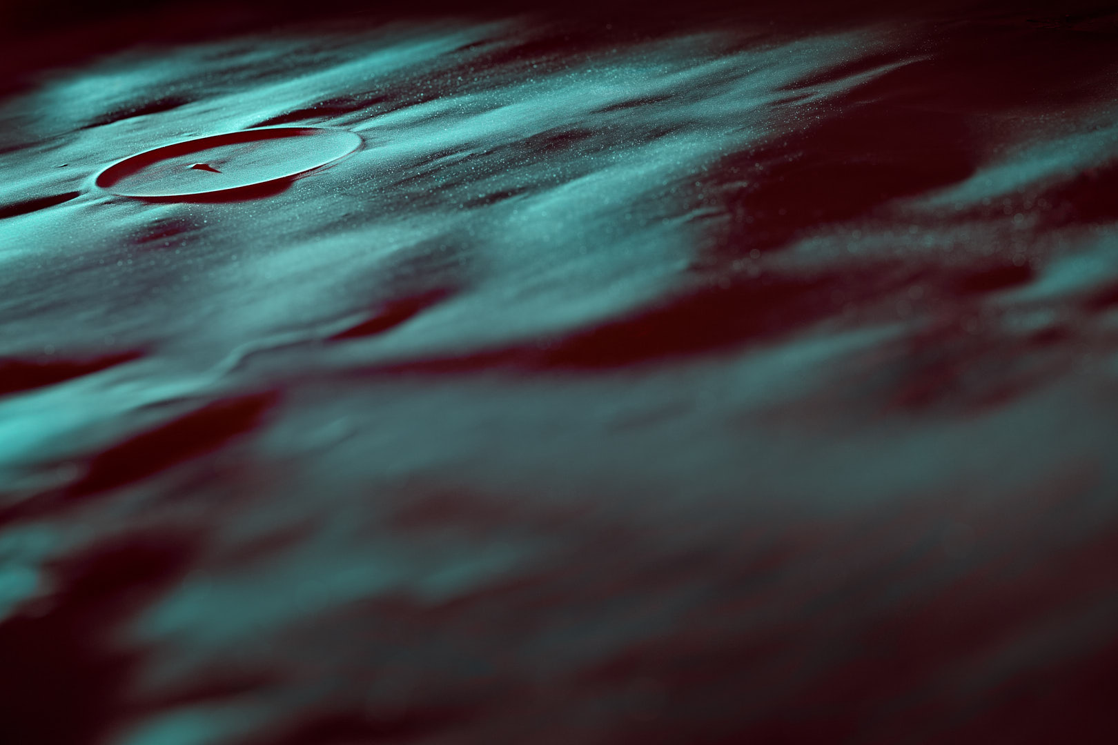 Abstract close-up viewed across a textured surface illuminated in turquoise and deep red, with a raised circular form near the upper left and shallow focus extending across the frame.