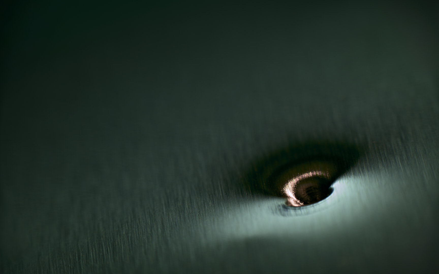 Dark green-gray abstract photograph of a softly textured surface with a small reflective circular depression and a pale band of light near the lower right.