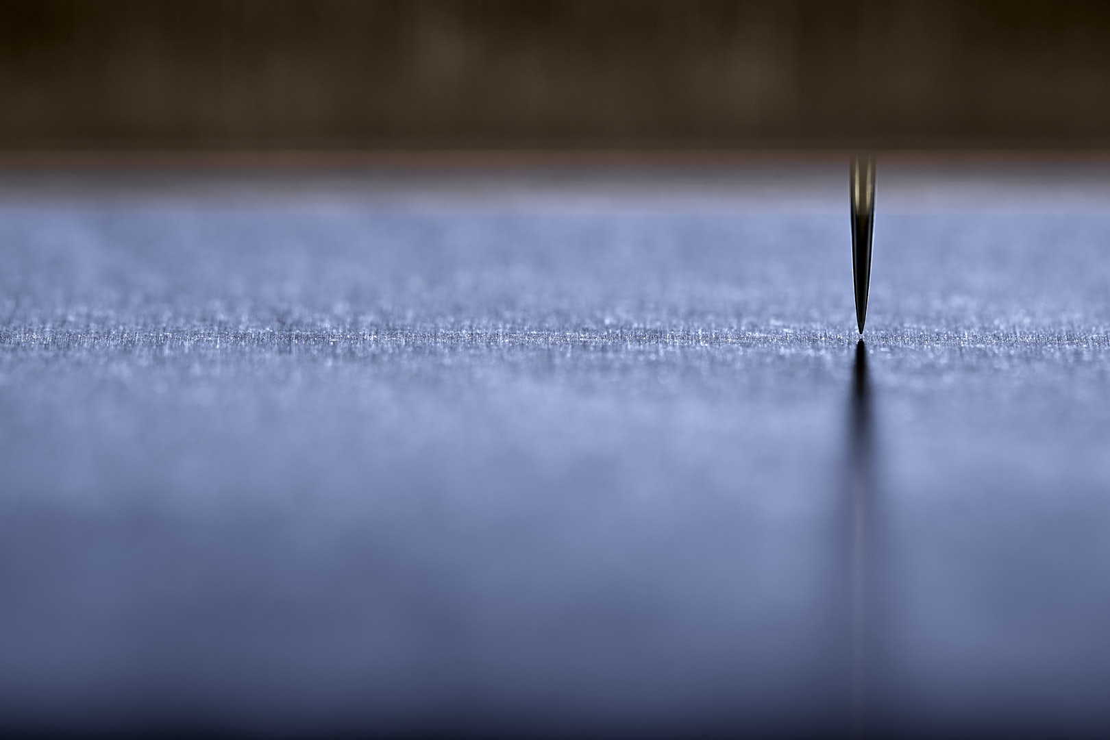 Abstract close-up of a pointed metal form suspended just above a pale blue-gray surface, with its reflection extending downward and broad softly focused horizontal fields surrounding it.