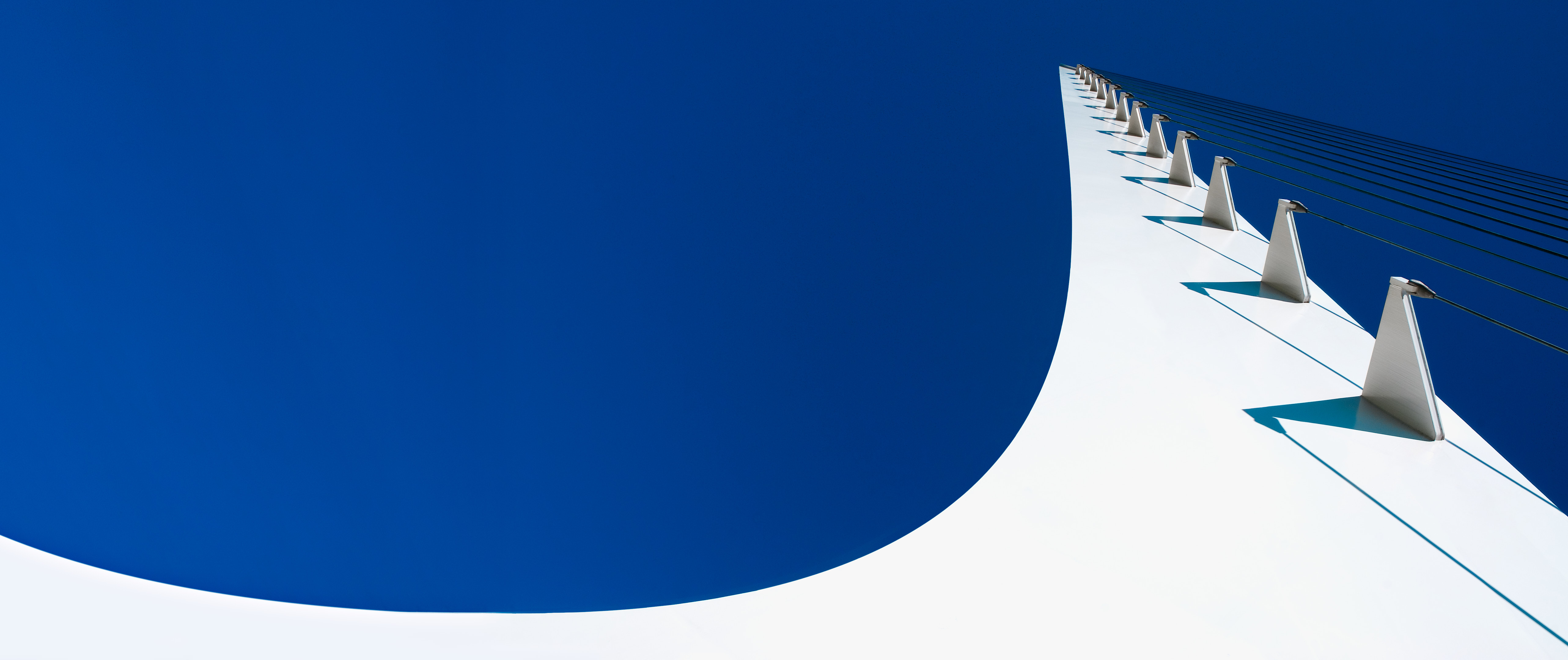 Upward view of the Sundial Bridge showing a sweeping white tower, repeating cable mounts and shadows, and taut cables against a deep blue sky.