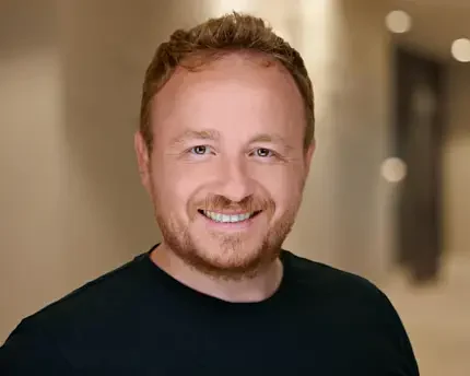 Corporate headshot of a male startup engineer in Palo Alto by Dean Birinyi, featuring a friendly smile and a black shirt against a blurred office background.