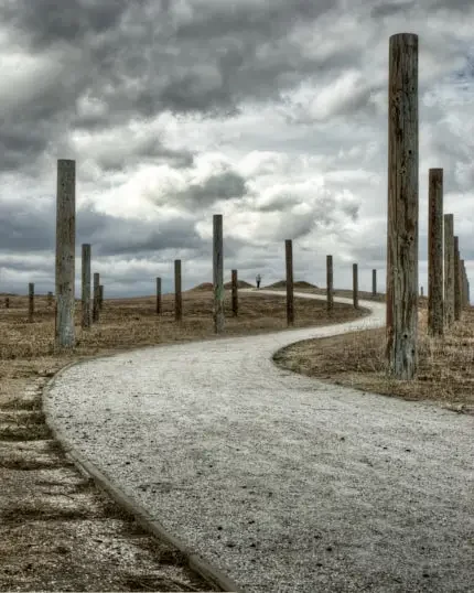 Pale curving path lined with tall wooden posts beneath a turbulent, cloud-filled sky.