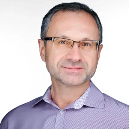 Professional headshot of a smiling man wearing glasses and a lavender button-down shirt against a white background.