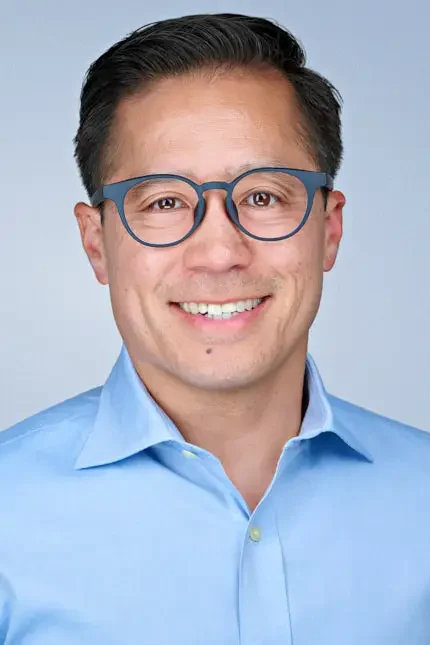 Professional corporate headshot of a smiling man wearing blue glasses and a light blue dress shirt against a light gray background.