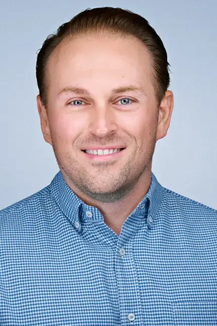 Professional headshot of a smiling man wearing a blue check button-down shirt against a light gray background.